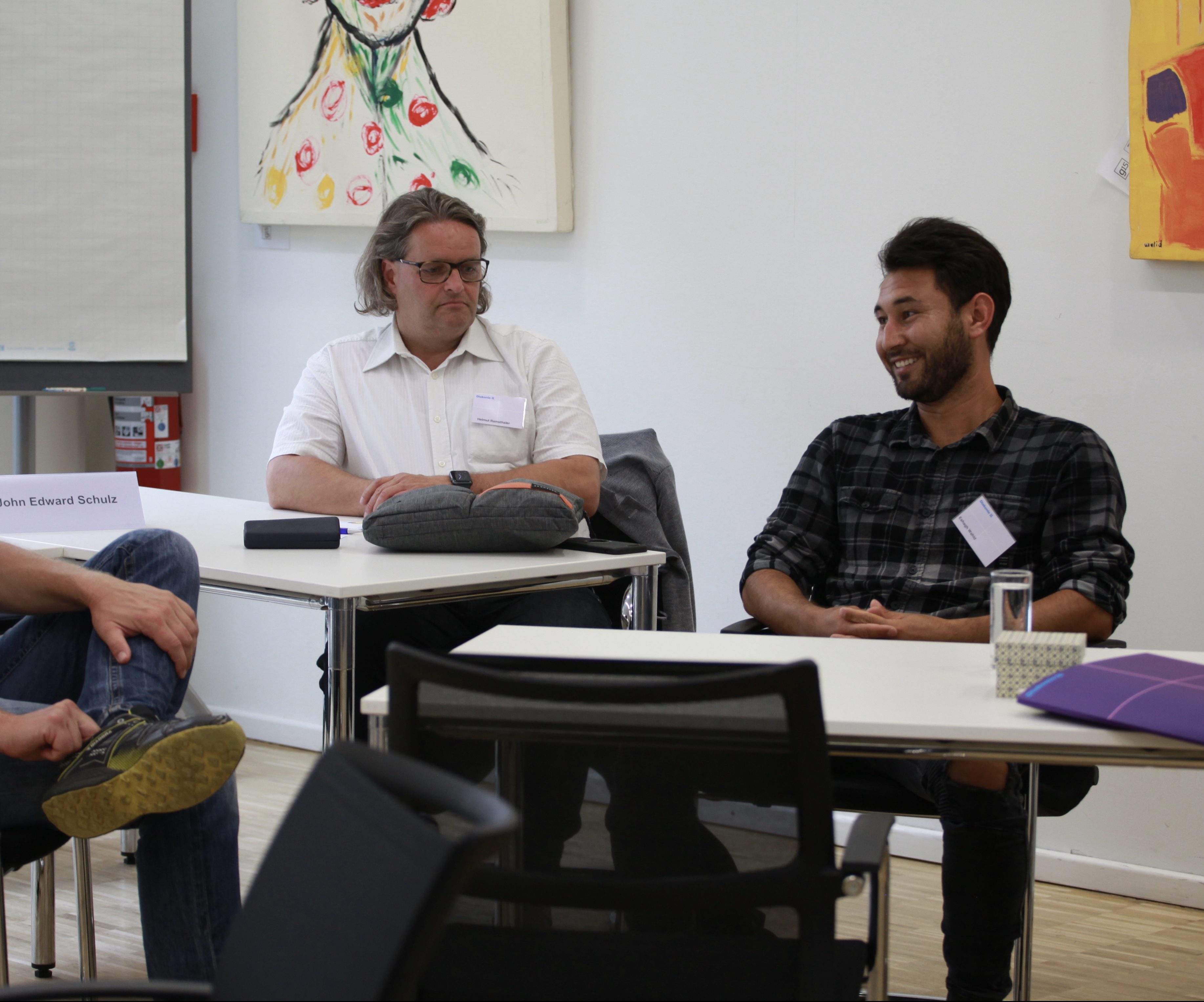 3 Männer im Seminarraum. Sie lächeln (größtenteils) während sie sich unterhalten.