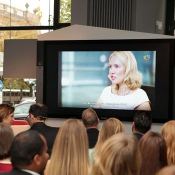 Eine junge, blonde Frau ist auf einem großen Bildschirm zu sehen, davor sind Menschen von hinten zu sehen, die in Stuhlreihen sitzen.