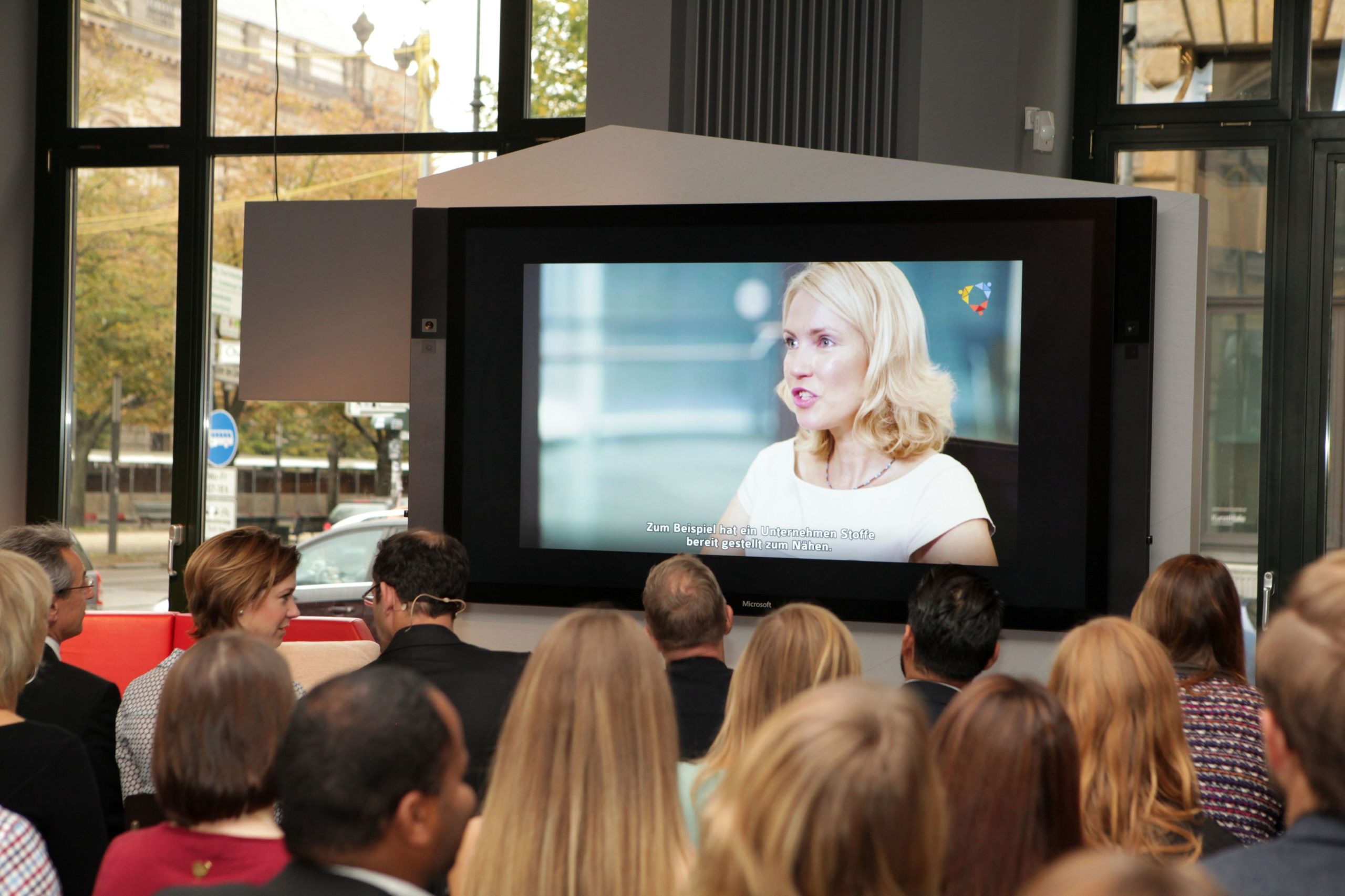 Eine junge, blonde Frau ist auf einem großen Bildschirm zu sehen, davor sind Menschen von hinten zu sehen, die in Stuhlreihen sitzen.