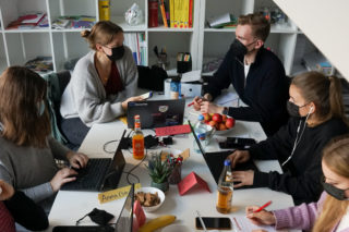 Menschen sitzen mit Masken auf dem Gesicht und Laptop vor sich auf einem Tisch