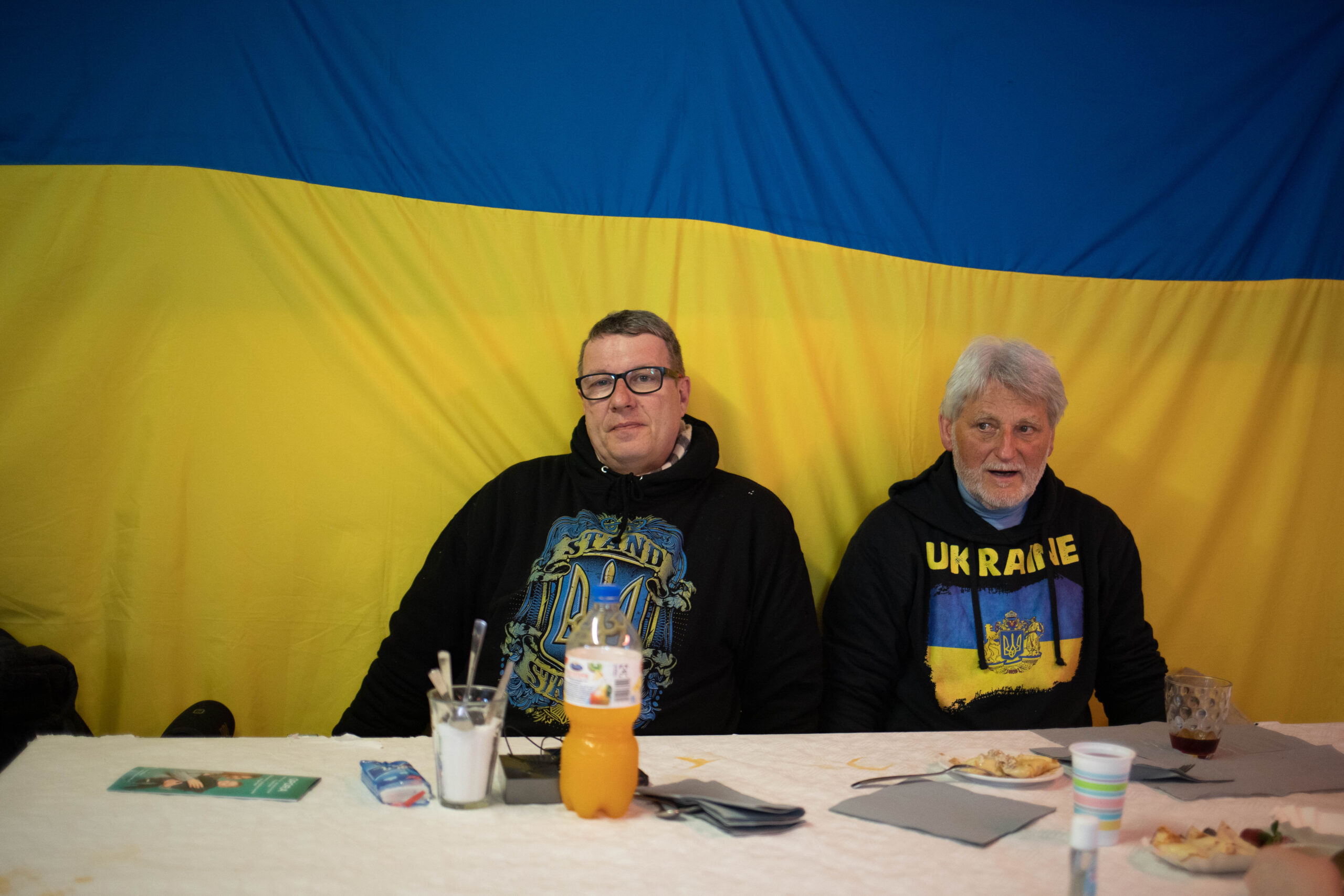 Es sind zwei Männer zu erkennen, die vor einer ukrainischen Flagge sitzen.