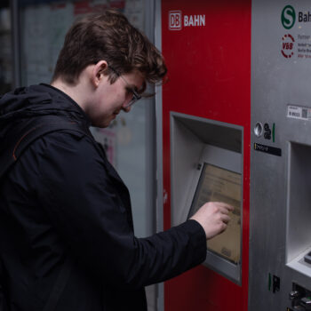 Michael steht an einem Ticketautomaten der Deutschen Bahn. Er trägt eine schwarze Jacke und einen Rucksack. Mit seiner rechten Hand tippt er auf dem Bildschirm des Automaten.
