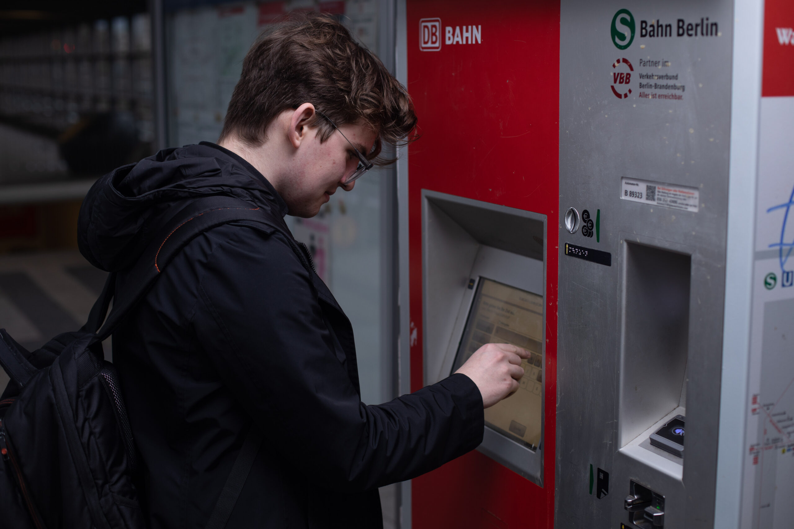 Michael steht an einem Ticketautomaten der Deutschen Bahn. Er trägt eine schwarze Jacke und einen Rucksack. Mit seiner rechten Hand tippt er auf dem Bildschirm des Automaten.