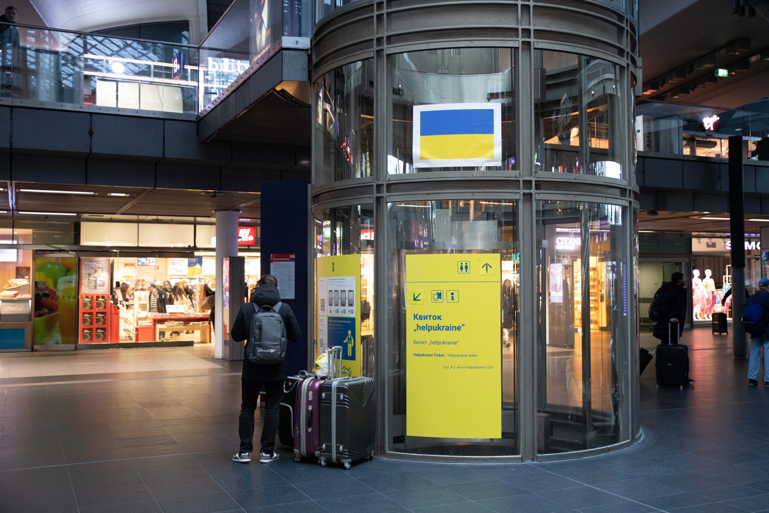 Ein gläserner Fahrstuhlschacht im Herzen des Berliner Hauptbahnhofs. Daran befestigt ist ein gelbes Plakat mit der Aufschrift “helpukraine” und einer Wegbeschreibung in kyrillischen Schriftzeichen.
