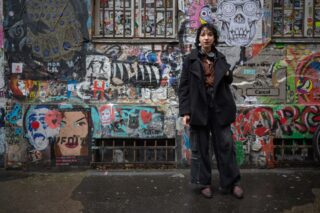 Auf dem Bild sieht man Nox vor einer grauen Betonwand stehen. Die Betonwand ist bemalt mit buntem Graffiti und beklebt mit einigen Stickern.Nox steht rechts im Bild, trägt eine schwarze Hose und einen schwarzen, langen Mantel. Um deren Hals hängen schwarze Kopfhörer. Nox hat schulterlange, lockige, schwarze Haare. Der Mantel ist auf und man sieht darunter eine braune Bluse.