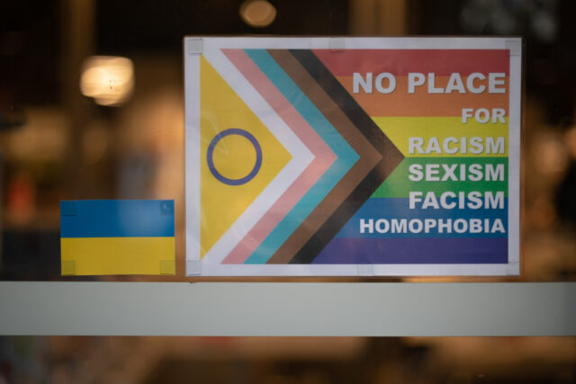Auf dem Bild sieht man aus der Nähe zwei Aufkleber auf einer Glasscheibe. Der Rechte Aufkleber ist Größer. Es ist eine LGBTQ Flagge auf der steht „No place for Racism, Sexism, Facism, Homophobia.“Links von der Flagge sieht man etwas kleiner die ukrainische Flagge.