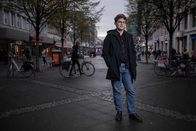 Michael steht in einer Fußgängerzone in Berlin-Charlottenburg. Er trägt eine schwarze Jacke und ein schwarzes Hemd mit einer blauen Jeans und schaut in die Kamera. Michael ist umgeben von Geschäften und Bäumen. Im Hintergrund sind verschwommen andere Menschen zu sehen.