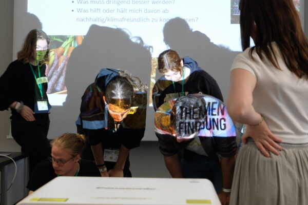 Das Foto zeigt fünf Teilnehmer*Innen über Materialien auf dem Boden gebeugt während der Themenfindung.