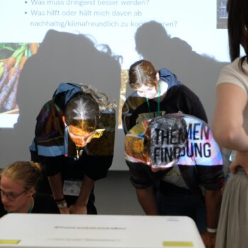 Das Foto zeigt vier Teilnehmer*Innen der Ernärhungssicherungs AG, die vor einem Beamer stehen und ein Plakat betrachten, das auf dem Boden liegt