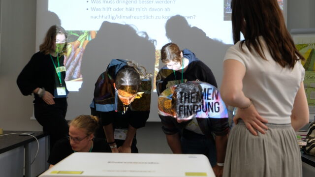 Das Foto zeigt vier Teilnehmer*Innen der Ernärhungssicherungs AG, die vor einem Beamer stehen und ein Plakat betrachten, das auf dem Boden liegt