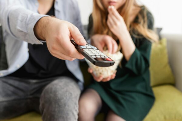 Zwei Menschen sitzen auf einer Couch, ein Mensch hält eine Fernbedienung in der Hand. Der andere Mensch isst Popcorn. 