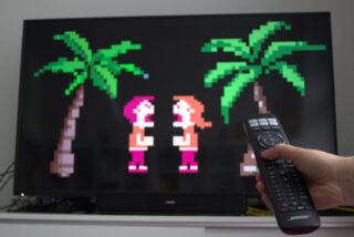 Eine Hand hält eine Fernbedienung. Im Hintergrund leicht unscharf ein Fernseher, auf dem Pixelfiguren im Streit zu sehen sind. 