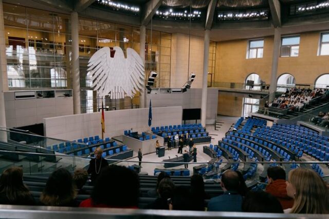 Heike Heubach steht am Redner:innenpult des Deutschen Bundestages, einige Personen stehen um sie herum. Das Bild wurde von der Zuschauertribüne aus gemacht.